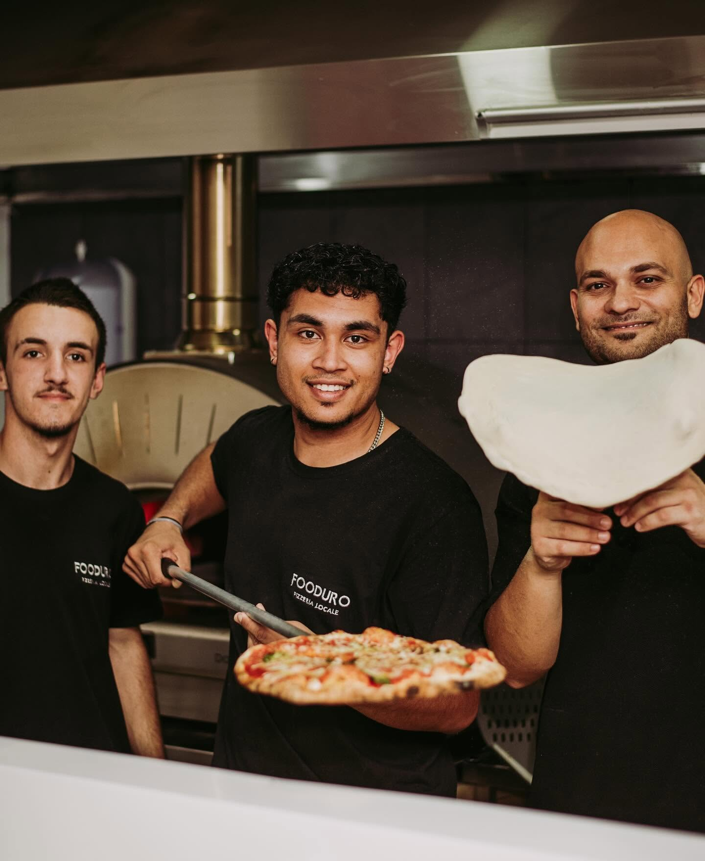 FOODURO pizzaiolo crew at the wood-fired oven holding a fresh pizza and dough